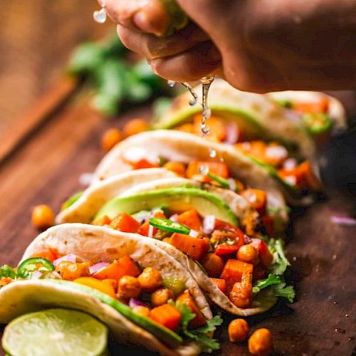 A hand squeezes lime over a row of tacos filled with chickpeas, avocado, and vegetables, garnished with cilantro, on a wooden surface.