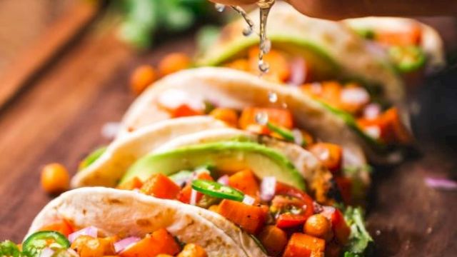 A hand squeezes lime over a row of tacos filled with chickpeas, avocado, and vegetables, garnished with cilantro, on a wooden surface.