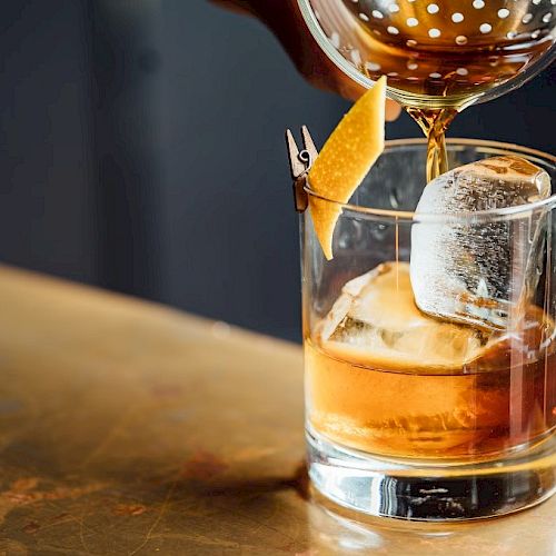 A cocktail is being poured into a glass with ice and a citrus peel garnish, sitting on a countertop.