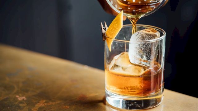 A cocktail is being poured into a glass with ice and a citrus peel garnish, sitting on a countertop.