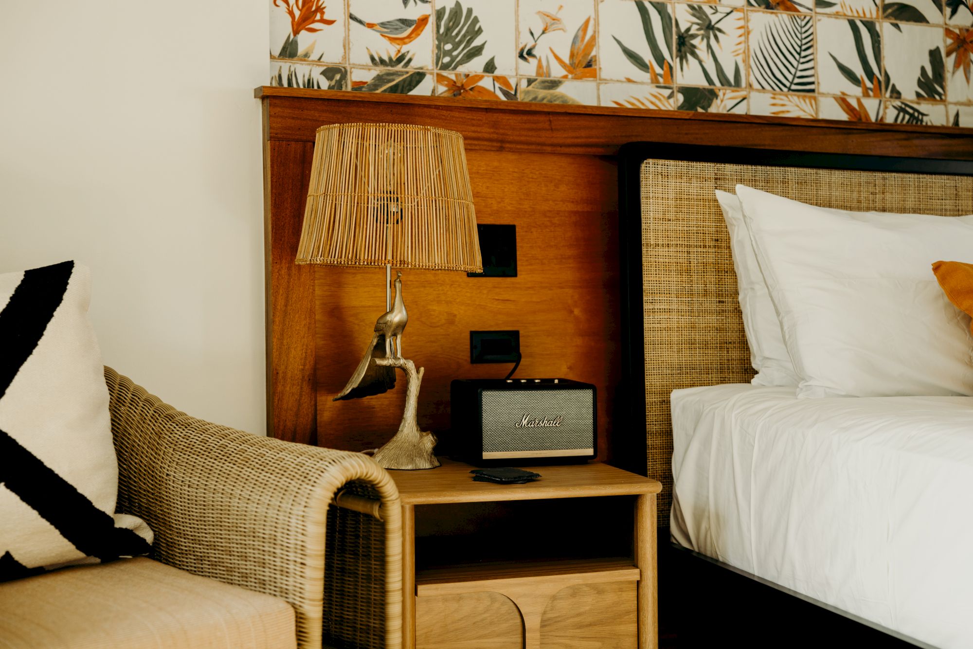 A cozy room with a wicker chair, a bedside table with a lamp and speaker, and a bed with patterned wallpaper in the background.