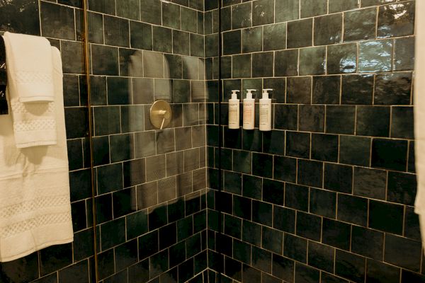 A modern shower with dark tiles, glass door, wooden stool, and bottles on the wall. Towels are hung nearby.