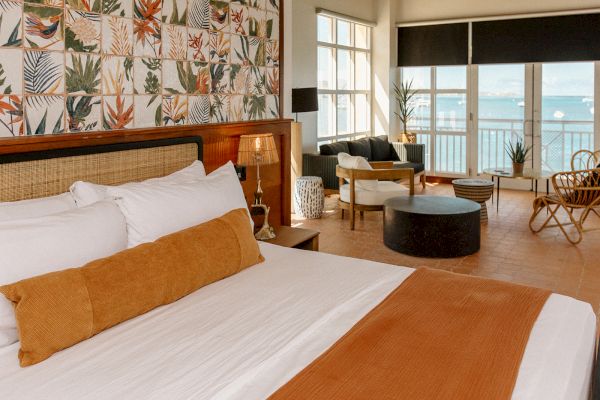 A cozy hotel room with a large bed, tropical decor, and a sitting area overlooks a balcony with a view of the sea.