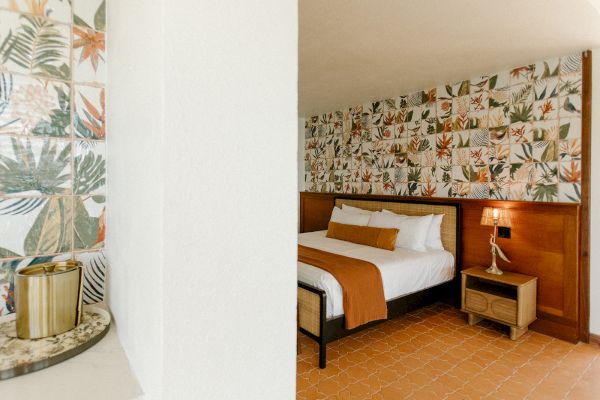 The image shows a cozy bedroom with a floral-themed wallpaper, a neatly made bed, and a bedside table with a lamp next to it.