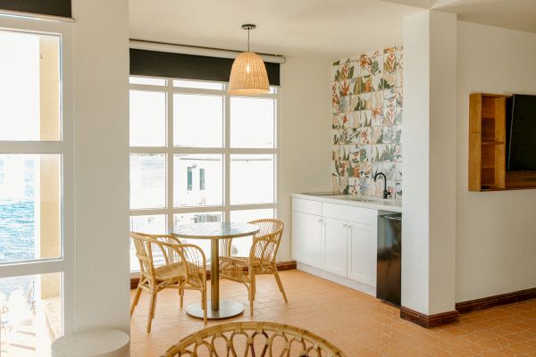 A bright room features a table with four wicker chairs, large windows, a small counter with tiles, and a hanging light fixture.