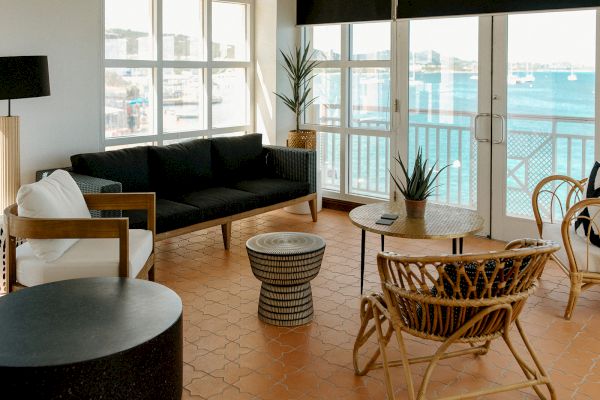 A stylish living room with wicker chairs, a black sofa, and ocean view windows. Coastal decor and a terracotta tile floor complete the scene.