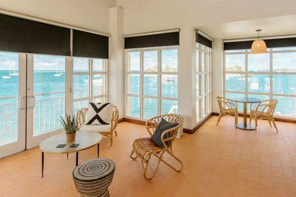 A sunlit room with wicker furniture, large windows, and ocean views. There's an inviting seating area and a small table by the window.