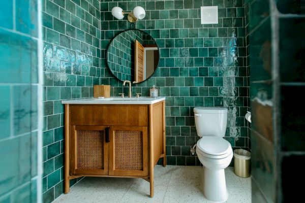 The image shows a bathroom with green tiled walls, a wooden vanity, a round mirror, a toilet, and a dual-light fixture above the mirror.