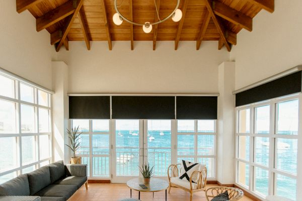 A coastal living room with large windows, a couch, chairs, and a view of the sea. The ceiling is wooden with a modern chandelier.