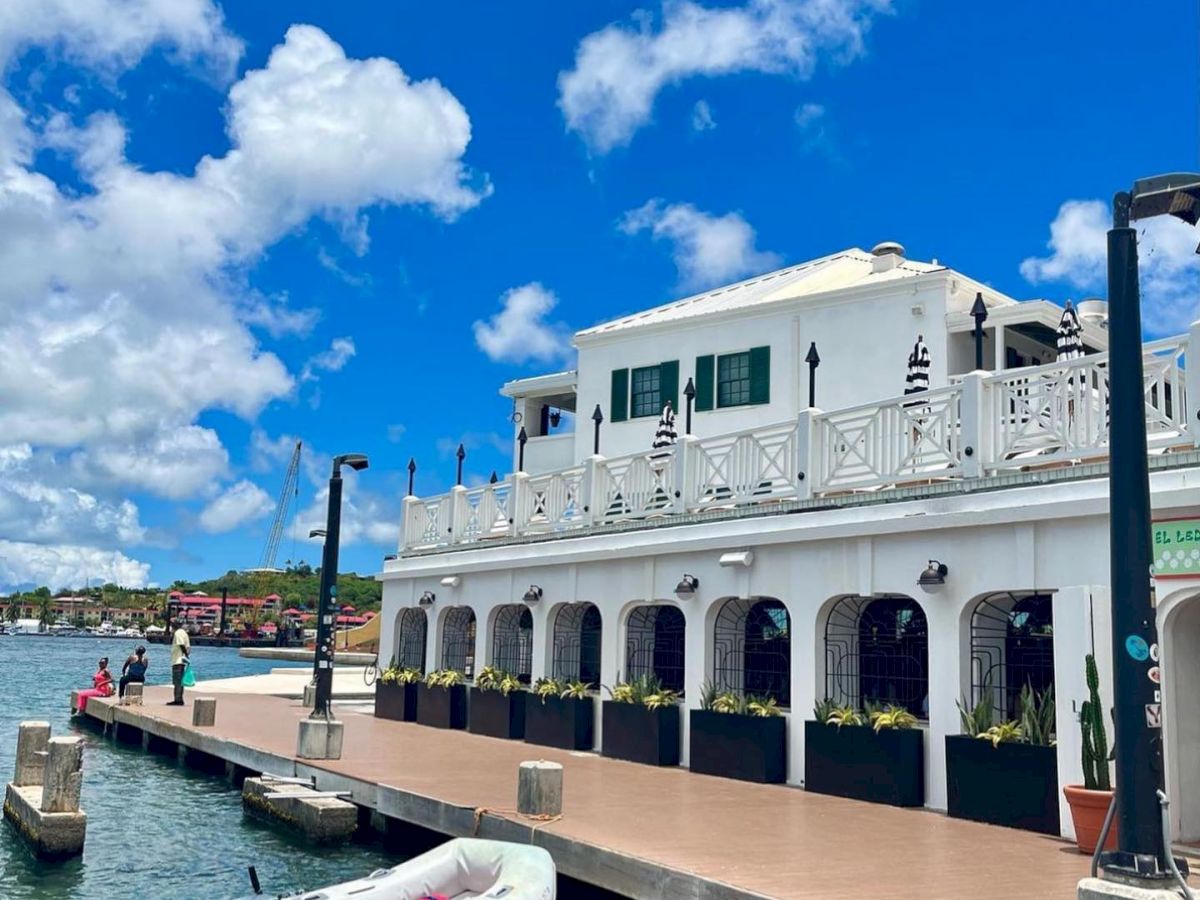 A white building with balconies overlooks a waterfront, with a docked inflatable boat and clear blue sky filled with fluffy clouds.