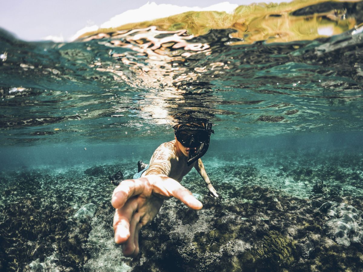 A person is snorkeling underwater, extending their arm toward the camera above a rocky seabed, with clear water and a shoreline visible.