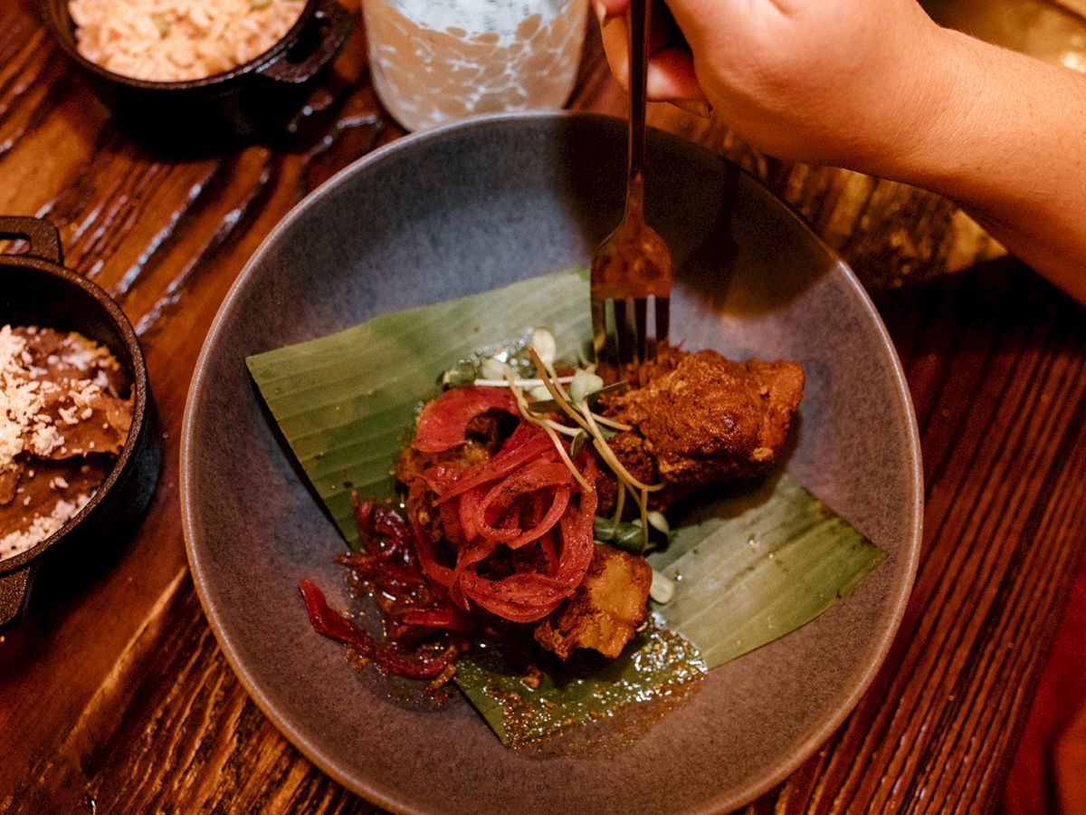 A person is holding a fork above a dish with meat, onions, and greens on a banana leaf. Sides of rice and beans are also on the table.