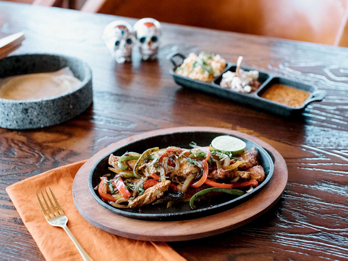 A platter of fajitas with lime, tortillas in a bowl, and side dishes on a wooden table, accented by orange napkin and fork.