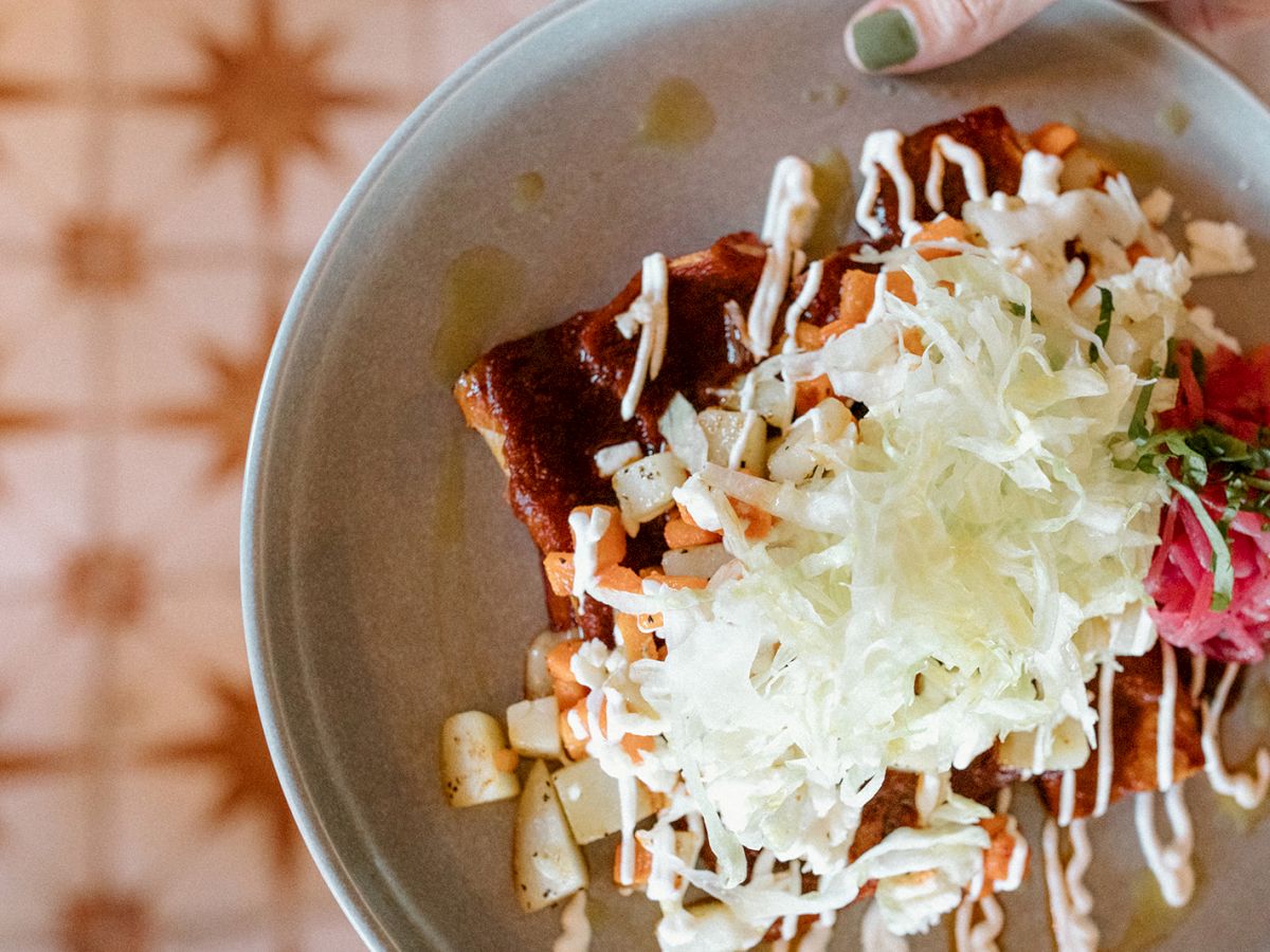 The image shows a plate of enchiladas topped with cheese, diced vegetables, creamy sauce, and a side of garnishes, served on a decorative table.