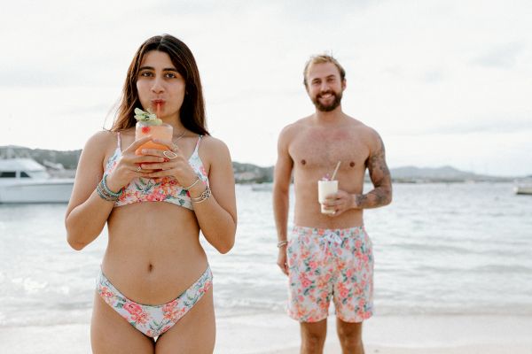 A woman and a man in matching floral swimwear enjoy drinks on a beach, with a boat in the background.