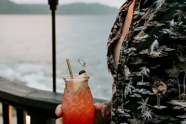 A person in a patterned shirt holds a tropical drink by the ocean, with a tiki torch lit in the background during sunset.