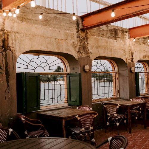 An outdoor seating area with wooden tables and chairs, arched windows, string lights, and a plant, creating a rustic ambiance.