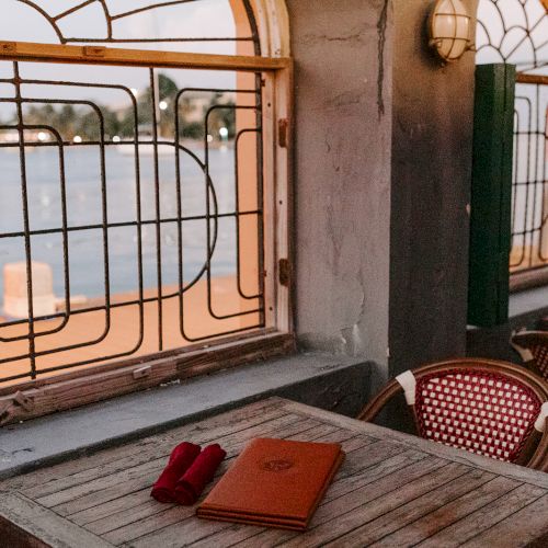 A cozy interior with wooden tables, chairs, decorative windows, and a view outside. A notebook and pen rest on a table.