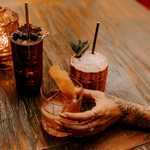 A tattooed hand holding a cocktail glass, with two tiki-style drinks and a decorative lantern on a wooden table.