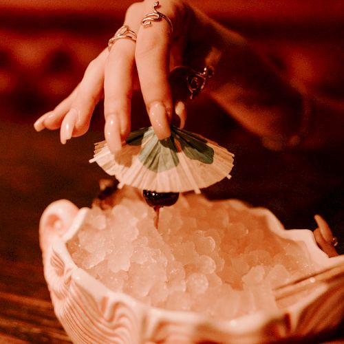 A hand with rings is holding a decorative paper umbrella over a bowl of ice cubes, creating a stylish and refreshing scene.