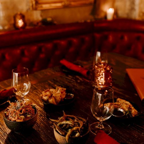 A cozy restaurant table with appetizers, wine glasses, red napkins, and warm candlelight ambiance in a dimly lit setting.