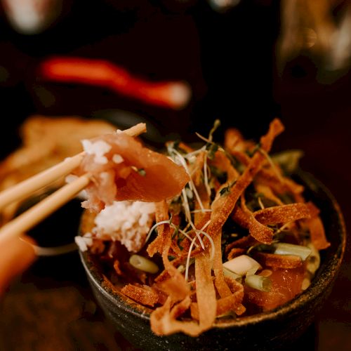 A bowl of food with chopsticks, featuring sliced meat, vegetables, and garnish, likely a dish of Asian cuisine.