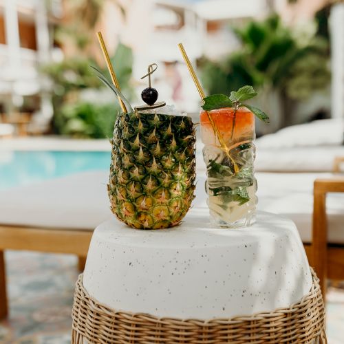 Two tropical drinks are placed on a round table by a poolside, one in a pineapple and the other in a glass with mint and a fruit garnish.