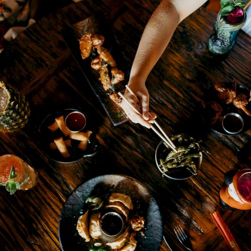 A top-down view of a table with various dishes and drinks, including skewers and garnished cocktails, and a hand serving food with tongs.