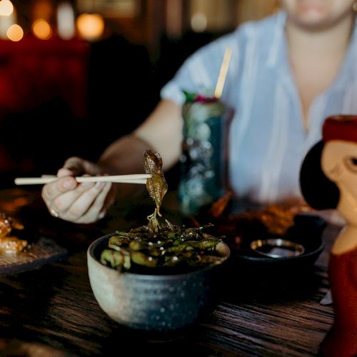 A person uses chopsticks to pick up food at a table with edamame, decorative cups, and dishes in a dimly lit setting.