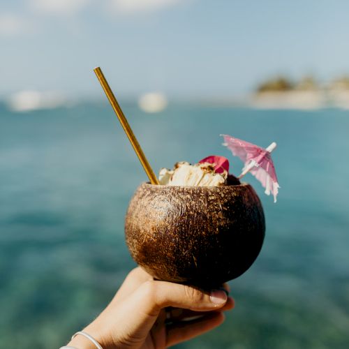 A hand holds a coconut drink with a straw and decorative umbrella by the sea, against a blurred ocean and sky background.