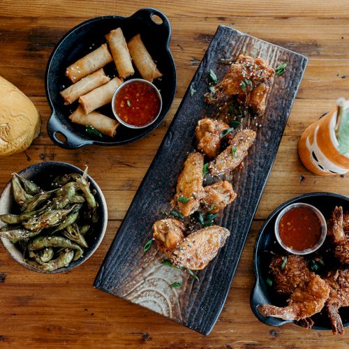 A wooden table with fried chicken, spring rolls, okra, shrimp, dipping sauces, and two drinks, including a mint garnish.