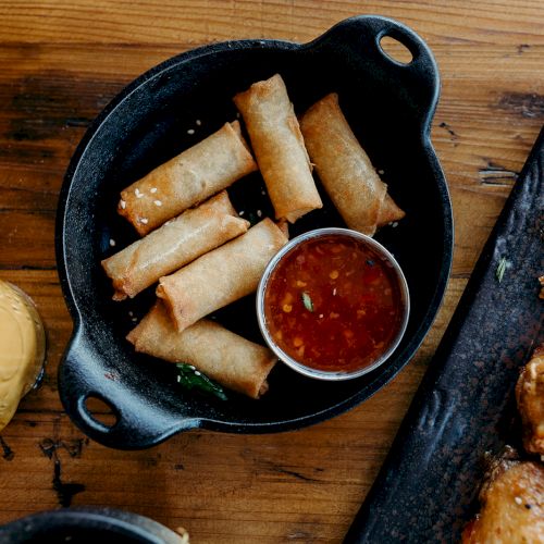 Spring rolls with dipping sauce are served in a black dish alongside some grilled meat on a wooden table.