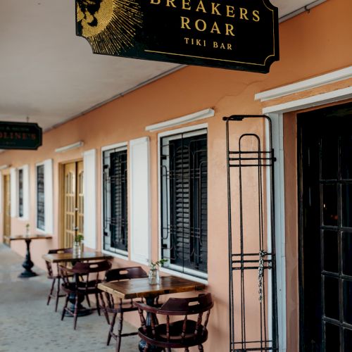 The image shows an outdoor seating area with small tables and chairs outside a bar named "Breakers Roar Tiki Bar" with a sign above.