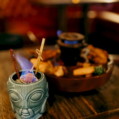 A tiki-style drink with fire and straws in a blue cup, alongside a plate of assorted food items on a wooden table.