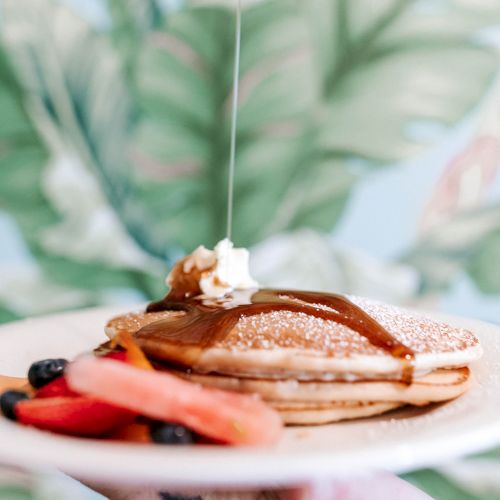 A plate of pancakes topped with butter and syrup, accompanied by fresh fruit slices, held against a leafy background.