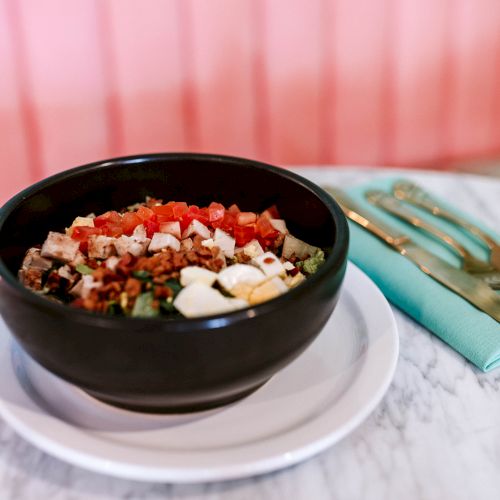 A bowl of colorful chopped salad with vegetables, cheese, and meat sits on a table, next to a napkin with silverware.