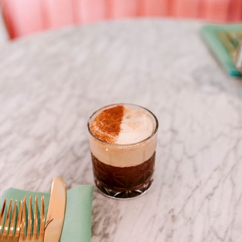 A glass with a layered coffee drink topped with foam sits on a marble table, next to gold cutlery on a green napkin.