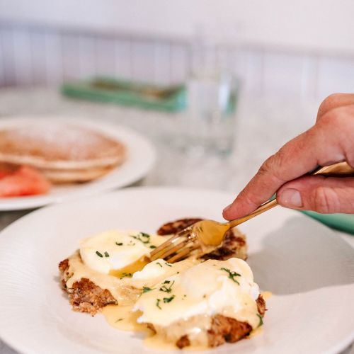 A person is using a fork to eat a dish with poached eggs and sauce on bread, with a side of pancakes in the background.