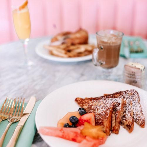 A breakfast spread featuring French toast with fruit, a drink with foam, and another plate in the background on a marble table setting.