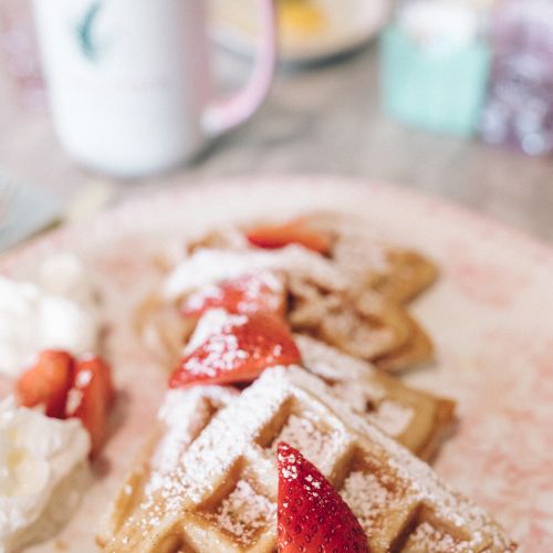 Waffles topped with strawberries and powdered sugar, served with whipped cream; a mug and more food items are seen in the background.