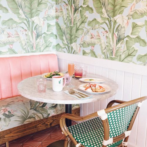 A stylish café corner with a pink booth, plant-patterned wallpaper, and a table set with breakfast items and drinks on a tiled floor.