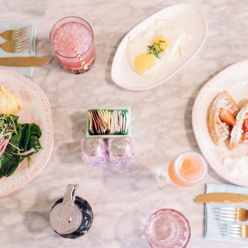 A tabletop with an omelette, greens, waffles with strawberries, butter on a dish, two drinks, cutlery, syrup, and a napkin holder.