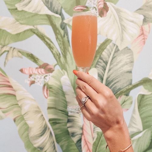 A hand holding a glass with an orange drink in front of a tropical leaf-patterned wallpaper.