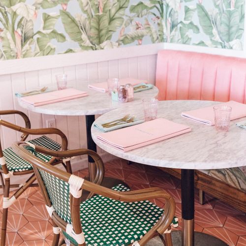 A cozy café with a marble table, pink chairs, and tropical wallpaper, set up with menus and glasses on a patterned tile floor.