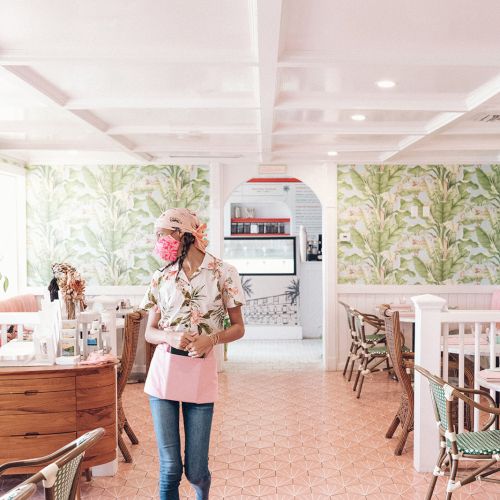 A woman walks through a brightly lit, cozy cafe with tropical wallpaper, wooden furniture, and a relaxed atmosphere.