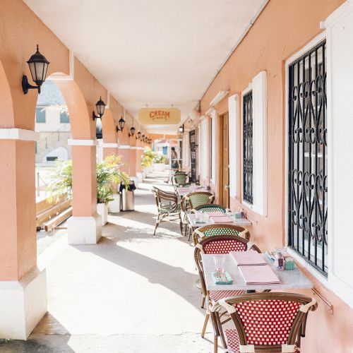 Outdoor cafe with tables and chairs along a covered walkway with pink walls and decorative arches, bathed in sunlight.