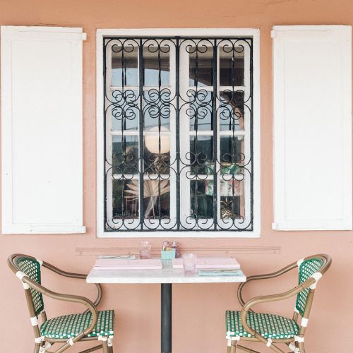 A small table with two chairs in front of a pink wall and a window with decorative bars.