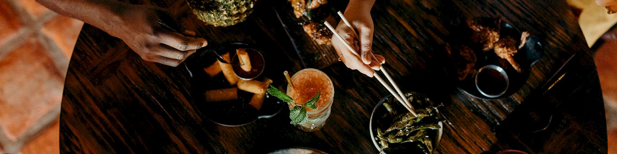 Aerial view of people dining, enjoying tropical drinks and a variety of dishes on a round wooden table.