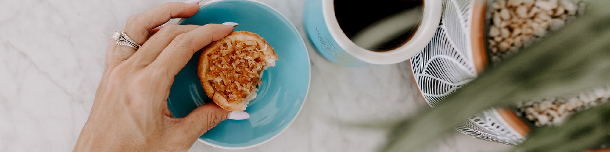 A hand holding a cookie over a blue plate, with a cup of coffee nearby on a marble surface next to a plant in a pot.