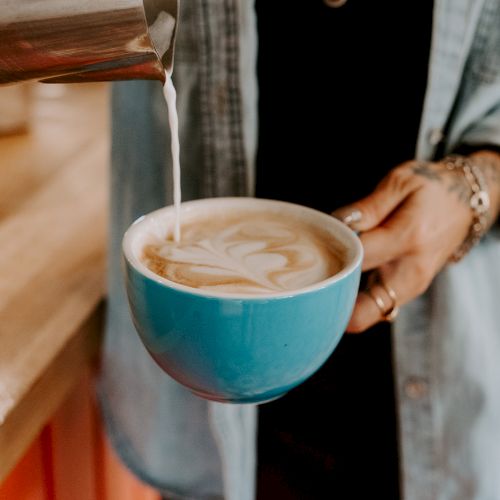 A person is pouring milk into a blue mug of coffee, creating latte art, with a wooden counter and casual clothing visible in the background.